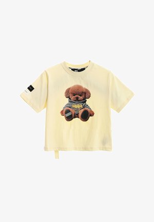 Camiseta de manga corta de color amarillo claro con un oso de peluche marrón con una sudadera con capucha gris estampada en la parte delantera y un parche en blanco y negro en la manga.