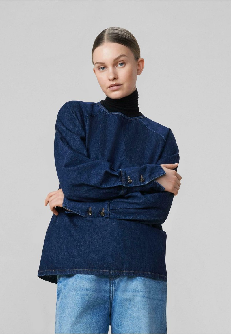 Top in denim blu scuro con scollatura rotonda, polsini con bottoni e vestibilità rilassata. Indossato sopra un dolcevita nero. Jeans in denim chiaro sotto.