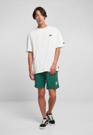 T-shirt bianca oversize in cotone con un piccolo logo, abbinata a pantaloni sportivi verdi con stampa grafica. Sneakers in bianco e nero.