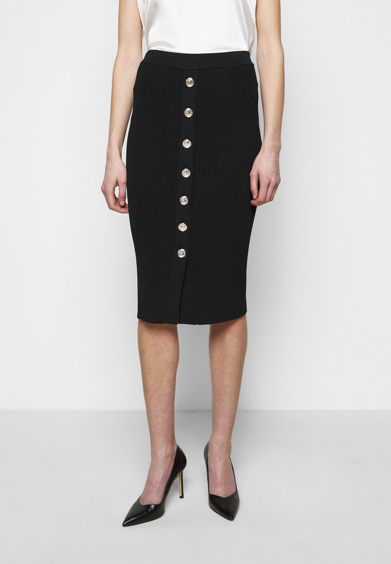 Femme portant une jupe crayon noire boutonnée avec des boutons argentés, associée à des escarpins noirs et un haut blanc sans manches.