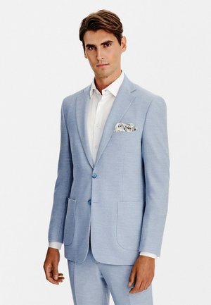 Mężczyzna w jasnoniebieskim garniturze, białej koszuli i wzorzystej poszetce, stojący na tle jednolitego białego tła.