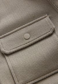 Gros plan sur un tissu beige texturé avec une poche à rabat fermée par un bouton rond beige.