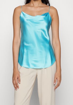 Kobieta ubrana w jedwabisty, jasnoniebieski, bezrękawnik z cienkimi ramiączkami oraz beżowe, luźne spodnie na białym tle.