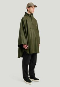 Olivi vihreä vedenpitävä poncho, jossa on korkea kaulus, etutasku vetoketjulla ja leveät hihat, yhdistettynä mustiin housuihin ja mustiin liukupohjaisiin kenkiin.