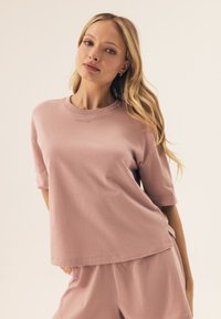 Jasnoróżowa, oversize'owa koszulka z krótkim rękawem, okrągłym dekoltem i subtelnym haftowanym logo na piersi. Gładki materiał bawełniany.