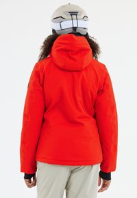 Person iført en lys rød hættejakke, beige bukser og en hvid hjelm, vist fra ryggen på en hvid baggrund.