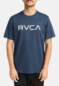 Hombre vestido con una camiseta de la marca RVCA de color azul marino con mangas cortas y pantalones negros, de pie frente a un fondo blanco liso.