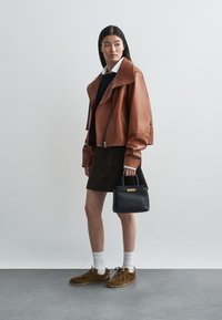 Jaqueta curta de couro castanho com colarinho oversized, combinada com um suéter preto, saia castanha e bolsa preta. A modelo usa tênis castanhos e meias brancas.