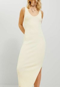 Frau trägt ein ärmelloses, geripptes, cremefarbenes Midikleid mit einem Seiten Schlitz und einer dünnen schwarzen Choker-Halskette.