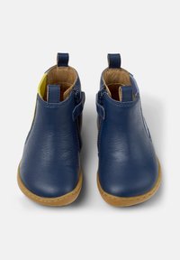 Une paire de petites chaussures en cuir marine pour tout-petits avec des semelles beige, une fermeture éclair sur le côté intérieur, et des poignées sur le talon et la languette, sur fond blanc.