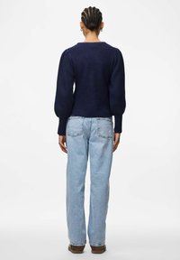 Tumši zila adīta džemperis ar puffētām garajām piedurknēm, apaļo apkakli un teksturētu apdari; kopā ar gaiši ziliem taisnajiem biksēm.