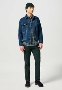 Chaqueta de mezclilla en azul oscuro, camisa de cuadros en verdes y azules, pantalones verdes, zapatos negros. Modelo de pie, wearing un gorro de lana verde.