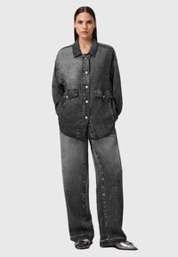 Donna in piedi con indosso una giacca di jeans nera oversize, lavata, con grandi bottoni e jeans a gamba larga abbinati, davanti a uno sfondo neutro.