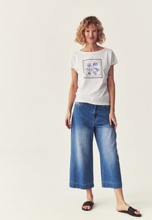 Frau mit lockigem Haar, die ein weißes T-Shirt mit Muschelprint, verkürzte weite blaue Jeans und schwarze Slide-Sandalen trägt, steht vor schlichter Hintergrund.