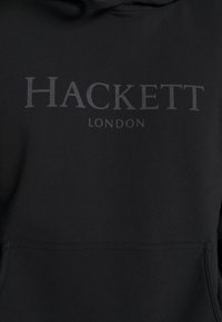 Černá mikina s kapucí, která má přední kapsu a velký šedý potisk s nápisem "HACKETT LONDON" přes hruď. Hladká textilie.