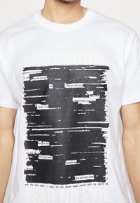 Hvid bomulds T-shirt med sort trykt tekst, delvist redigeret med horisontale sorte streger. Inkluderer "FORTROLIG" og "FRYR INTET ONDT" nederst.