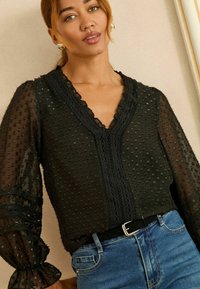 Čierna priehľadná blúzka s textúrovanými bodkami a čipkovým lemovaním pozdĺž V-výstrihu, s vyp puffed rukávmi a zložitými šitými detailmi.