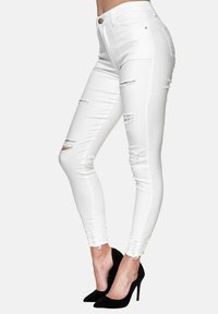 Jeans ajustados de mezclilla blanca con detalles desgastados, dobladillo deshilachado y adornos en el tobillo. Combinados con tacones de aguja negros.