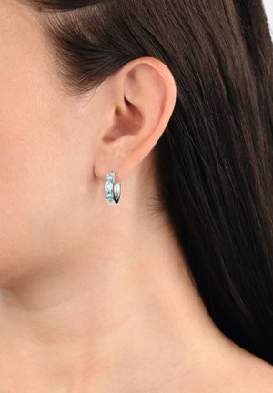 Silberne Creolen mit hellblauen Edelsteinen, die am linken Ohr einer Frau getragen werden, Seitensicht mit dunkelbraunem Haar, das hinter dem Ohr gesteckt ist.