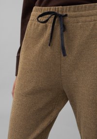 Bruine geblokte broek met een zachte textuur, voorzien van een tailleband met trekkoord in marineblauw. Close-up van het stofpatroon en de details van de tailleband.