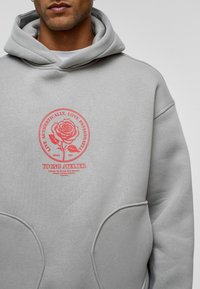 Grijze hoodie met een grote, rode cirkelvormige graphic met een rozenontwerp en de tekst "LEEF AUTHENTIEK, LEEF MET PASSIE." Grote voorzakken.