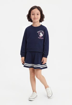 Lächelndes Kind mit kurzen dunklen Haaren trägt ein marineblaues Minnie-Maus-Sweatshirt, einen passenden Rock mit weißen Streifen und weiße Turnschuhe, steht aufrecht.