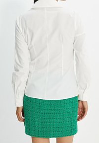 Camicia bianca a maniche lunghe con colletto classico, abbinata a una gonna verde testurizzata con un motivo intrecciato e taglio dritto.