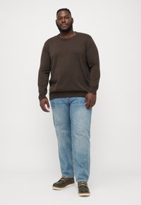 Brun crewneck genser, falmede blå rette jeans og mørke grønne støvler med beige såler, som står mot en hvit bakgrunn.