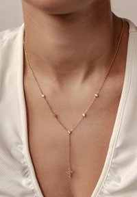 Collar de cadena dorada con pequeños acentos redondos y un colgante en forma de Y que presenta un colgante de cruz adornado con piedras transparentes. Fondo texturizado.