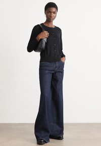 Svart knappet cardigan med rund hals, kombinert med utslåtte mørke denimjeans. Modellen holder en svart håndveske og står foran en nøytral bakgrunn.