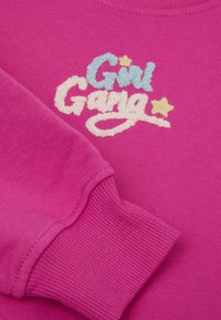 Růžová mikina se vyšívaným bílým, světle modrým a žlutým textem, který říká "Girl Gang". Žebrované manžety a měkká textura látky.