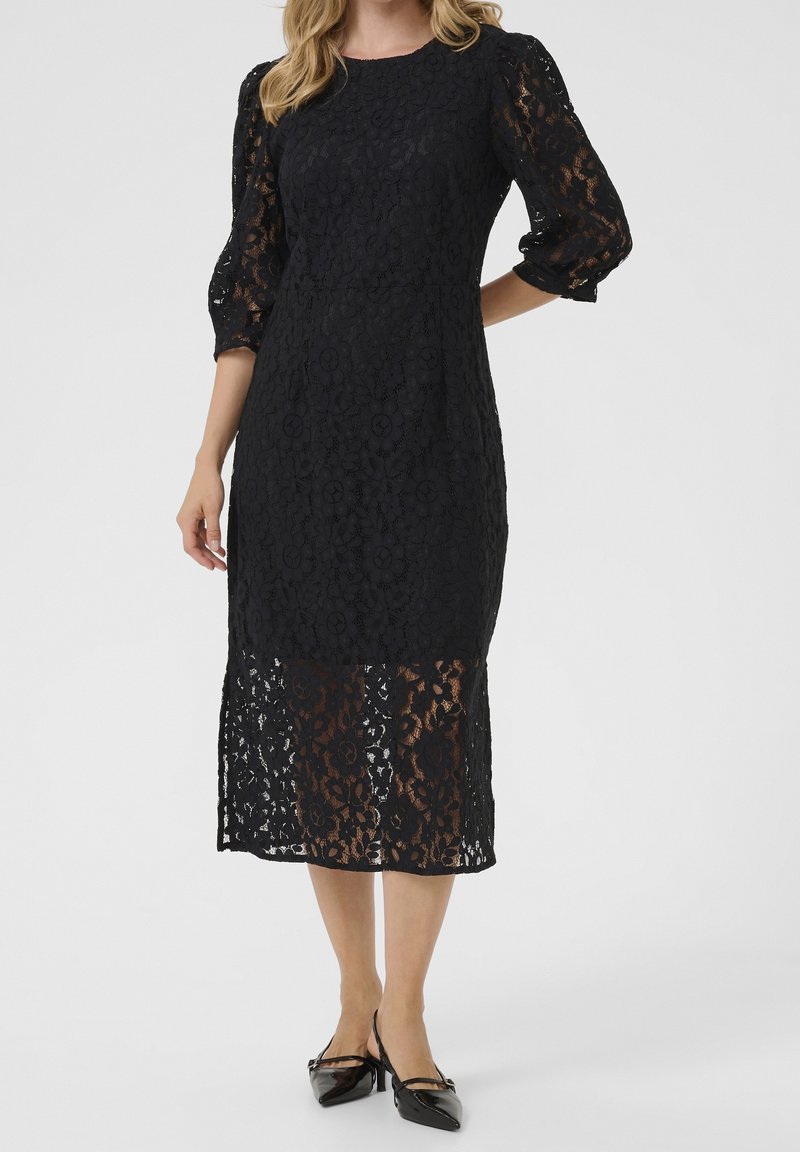 Femme portant une robe midi en dentelle noire avec des manches trois-quarts bouffantes et des talons pointus noirs sur fond blanc.
