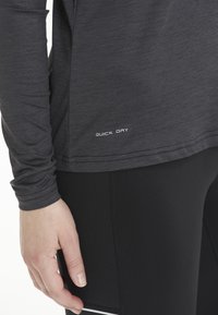 Langärmliges schwarzes Funktionsshirt mit strukturierter Oberfläche. Etikett mit "QUICK DRY" in Weiß eingestickt. Getragen mit schwarzen Leggings.