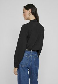 Musta pitkähihaine paita frillikauluksella, yhdistettynä korkeavyötäröisiin sinisiin farkkuihin, joissa on klassinen viiden taskun design.