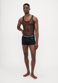 Schwarzes Netz-Tanktop mit dünnen Trägern, kombiniert mit schwarzen Shorts mit einem gemusterten Bund. Glatte Textur, figurbetontes Design.