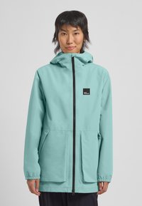 Veste imperméable bleu clair avec capuche, fermeture éclair avant et deux poches inclinées. Dotée de poignets élastiques et d'un petit logo noir sur la poitrine.