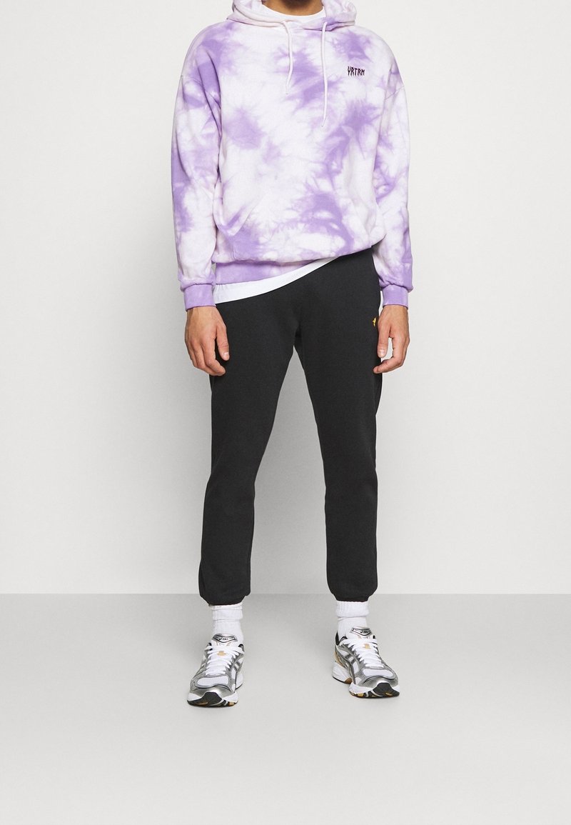 Homme portant un sweat à capuche tie-dye violet et blanc, un pantalon de jogging noir, des chaussettes blanches et des baskets gris-blanc, debout contre un mur uni.