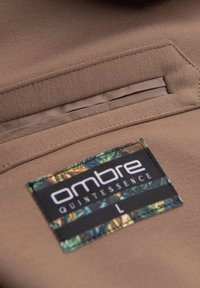 Brauner Stoff mit glatter Textur, versehen mit einem Patch-Label mit "ombre QUINTESSENCE" und einem blätterartigen Muster in Grün und Gelb. Größe: L.