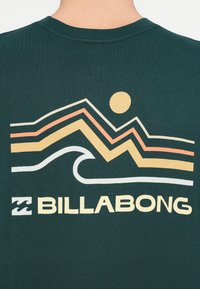 Σκούρο πράσινο T-shirt με γραφικό που απεικονίζει πολύχρωμες ζιγκ-ζαγκ γραμμές, έναν ήλιο και ένα κύμα, με το κείμενο "BILLABONG" σε έντονη λευκή γραμματοσειρά.