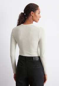 Langermet lys beige topp med ribbestruktur, rund halslinning og tettsittende design, kombinert med høytlivs svarte jeans og et logoparche.