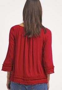 Röd, texturerad blus med trekvartslång ärm. Har spetsdetaljer vid ärmsluten och fållen. Avslappnad passform med rynkad detalj baktill.
