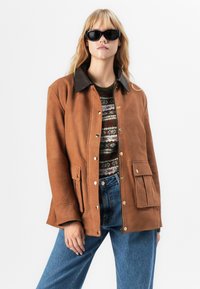 Una donna con capelli biondi indossa occhiali da sole neri, una giacca di camoscio marrone con tasche frontali, un maglione con un motivo e jeans di denim blu.