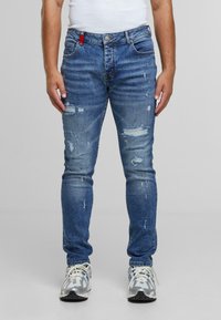 Moški nosi prilegajoče se modre uničene kavbojke z madeži od barve in bele superge, stoji ob ozadju brez vzorca.