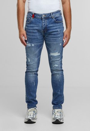 Moški nosi prilegajoče se modre uničene kavbojke z madeži od barve in bele superge, stoji ob ozadju brez vzorca.