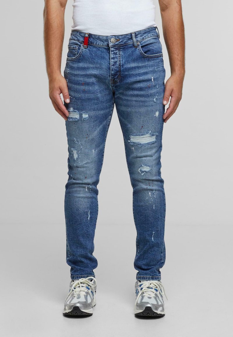 Moški nosi prilegajoče se modre uničene kavbojke z madeži od barve in bele superge, stoji ob ozadju brez vzorca.