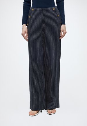 Pantaloni bleumarin cu dungi fine și croială largă, cu nasturi aurii pe părți, purtați cu top negru și tocuri albe decupate în față.