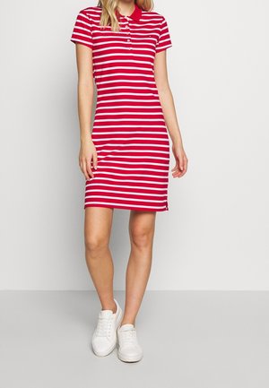 Vrouw draagt een rode en witte polo-jurk met horizontale strepen en witte sneakers, staand tegen een effen lichte achtergrond.