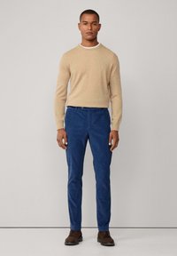 Maglione beige in maglia con scollo rotondo, abbinato a pantaloni in corduroy blu. Scarpe marroni completano l'outfit. Design semplice e contemporaneo.