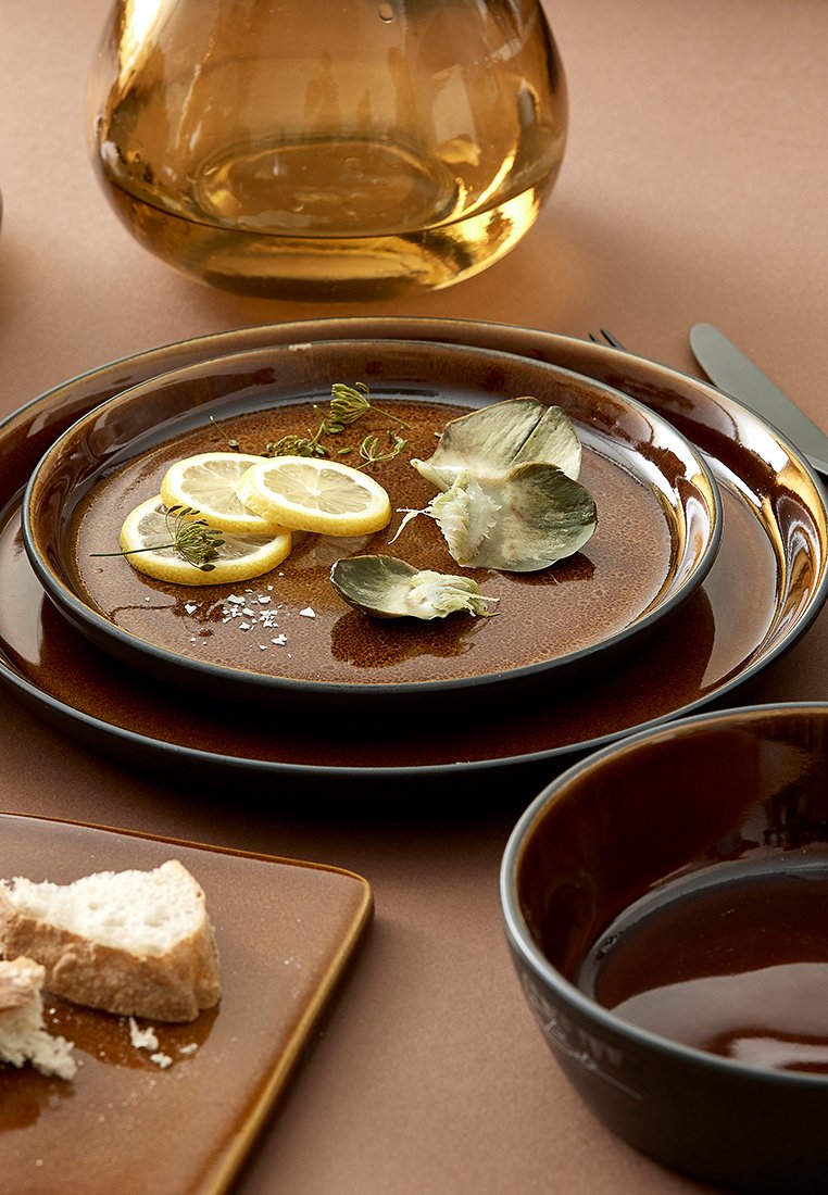 Piatti di ceramica marrone presentano fette di limone e carciofo, guarniti con erbe, accanto a una ciotola marrone e pane tostato, su una superficie neutra.