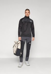 Crna jakna na patent s sivim detaljima, uparena sa sivim i crnim hlačama. Model drži bež i crni ruksak. Svijetlosive cipele.